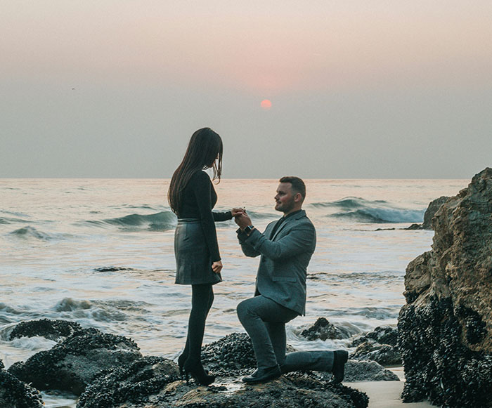 Pareja en la playa durante la puesta de sol con un hombre arrodillado para proponerle matrimonio en una sesión de fotos de compromiso por parte de un fotógrafo.